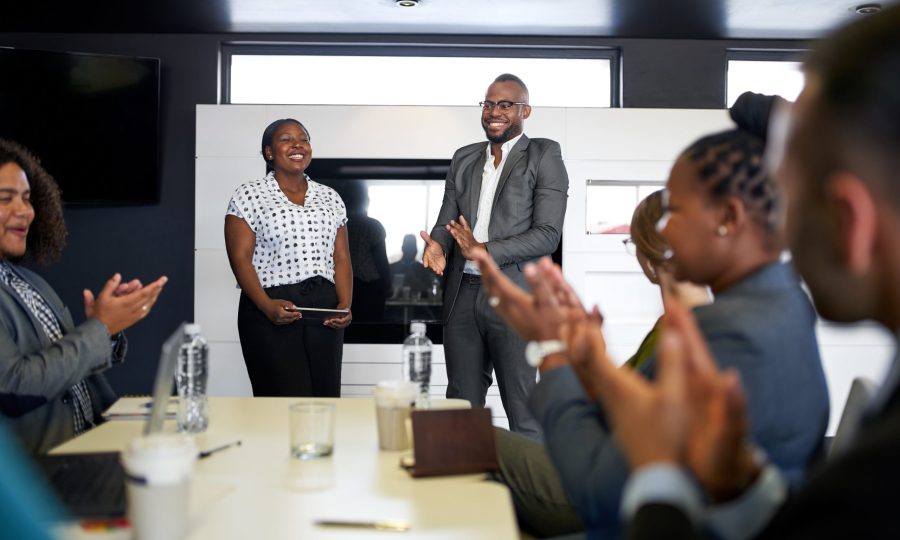 Group of mixed race colleagues in modern meeting room with laptop computer encouraging two attractive African American business professionals leading a collaborative discussion