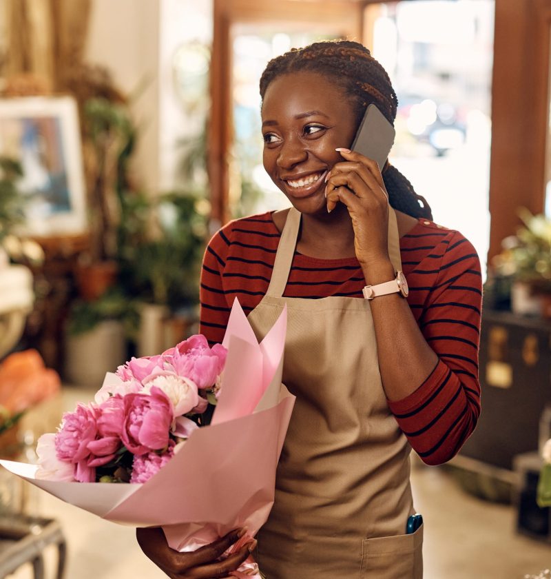 Happy African American florist holding bouquet of flowers and talking on cell phone at flower shop.